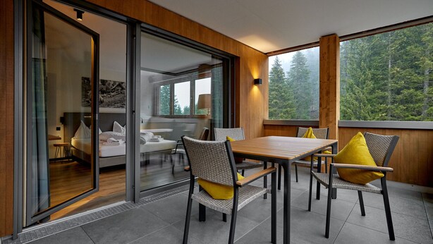 Un interior moderno con zona de comedor que incluye una mesa y sillas de madera, un ventanal de vidrio que da a un dormitorio y vistas a un bosque a través de amplios ventanales.