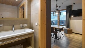 Un baño moderno con un lavabo grande, un espejo y detalles de madera. Hay una zona de comedor con una mesa y sillas, y una vista de la montaña a través de un ventanal grande.
