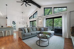 A spacious living area with a grey sectional sofa, a round coffee table, and large windows offering a view of trees. - Poconos Chatham Continental - Hot Tub & Fire Pit! (Bushkill)
