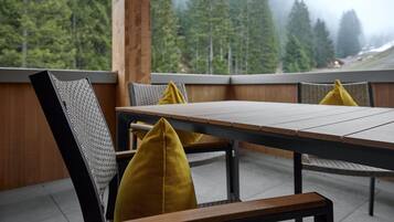 A wooden table with chairs, each with a yellow cushion, set against a backdrop of a forested mountain range.