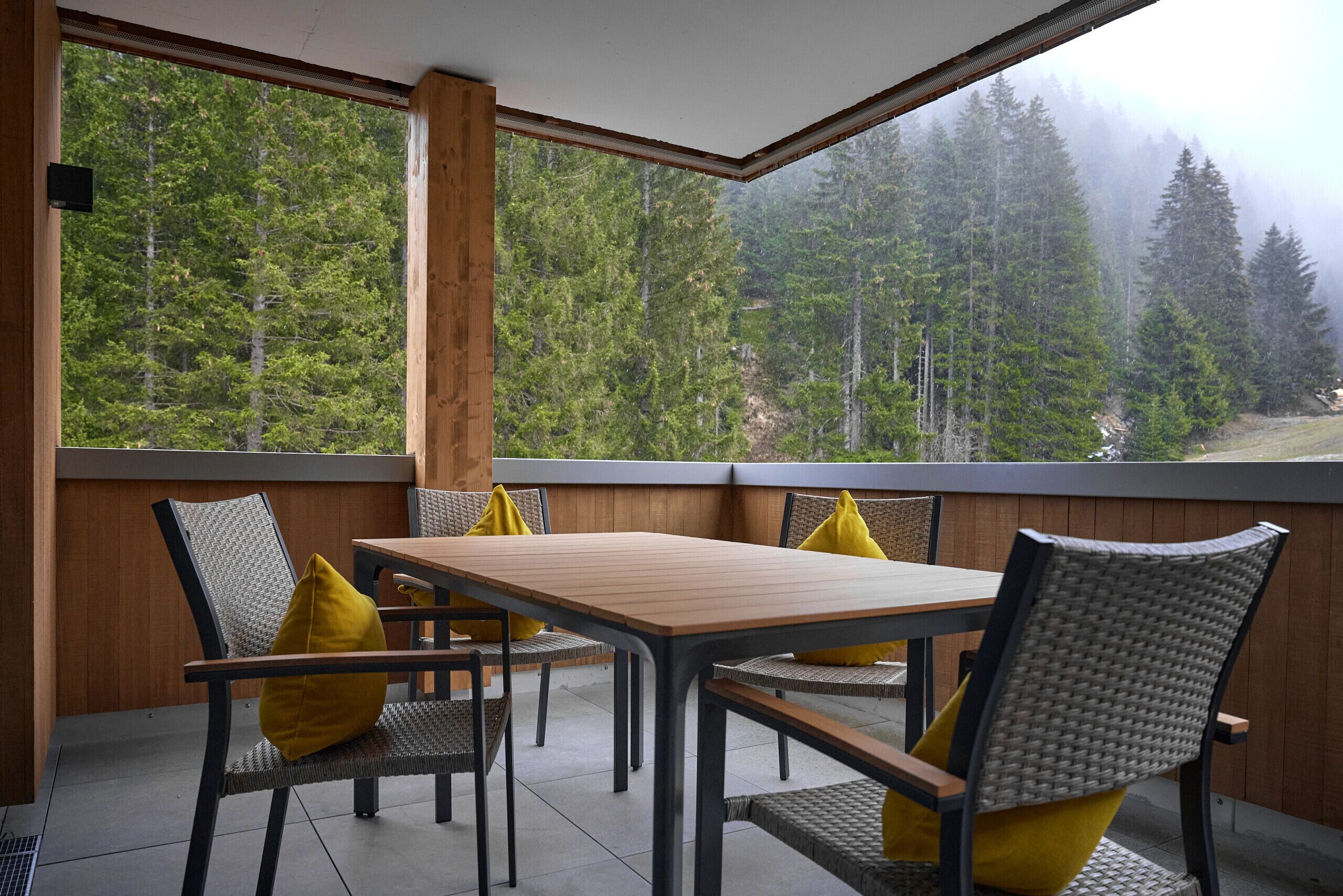 Un balcon avec une table et des chaises en bois, donnant sur une zone boisée grâce à de grandes fenêtres.