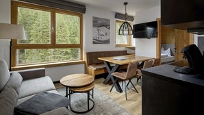 Ein modernes Zimmer mit einer Couch, einem Esstisch mit Stühlen, einer Bank, einem an der Wand befestigten Fernseher und großen Fenstern mit Blick auf einen Wald.