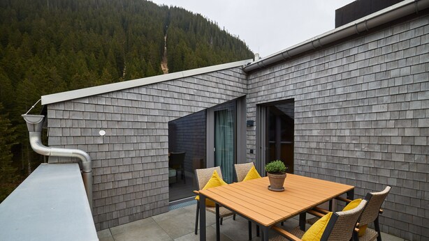 A modern outdoor patio with a wooden table and chairs, a potted plant, and a view of a forested mountain. - 59217 Wanderfalke 217 T (Gargellen)