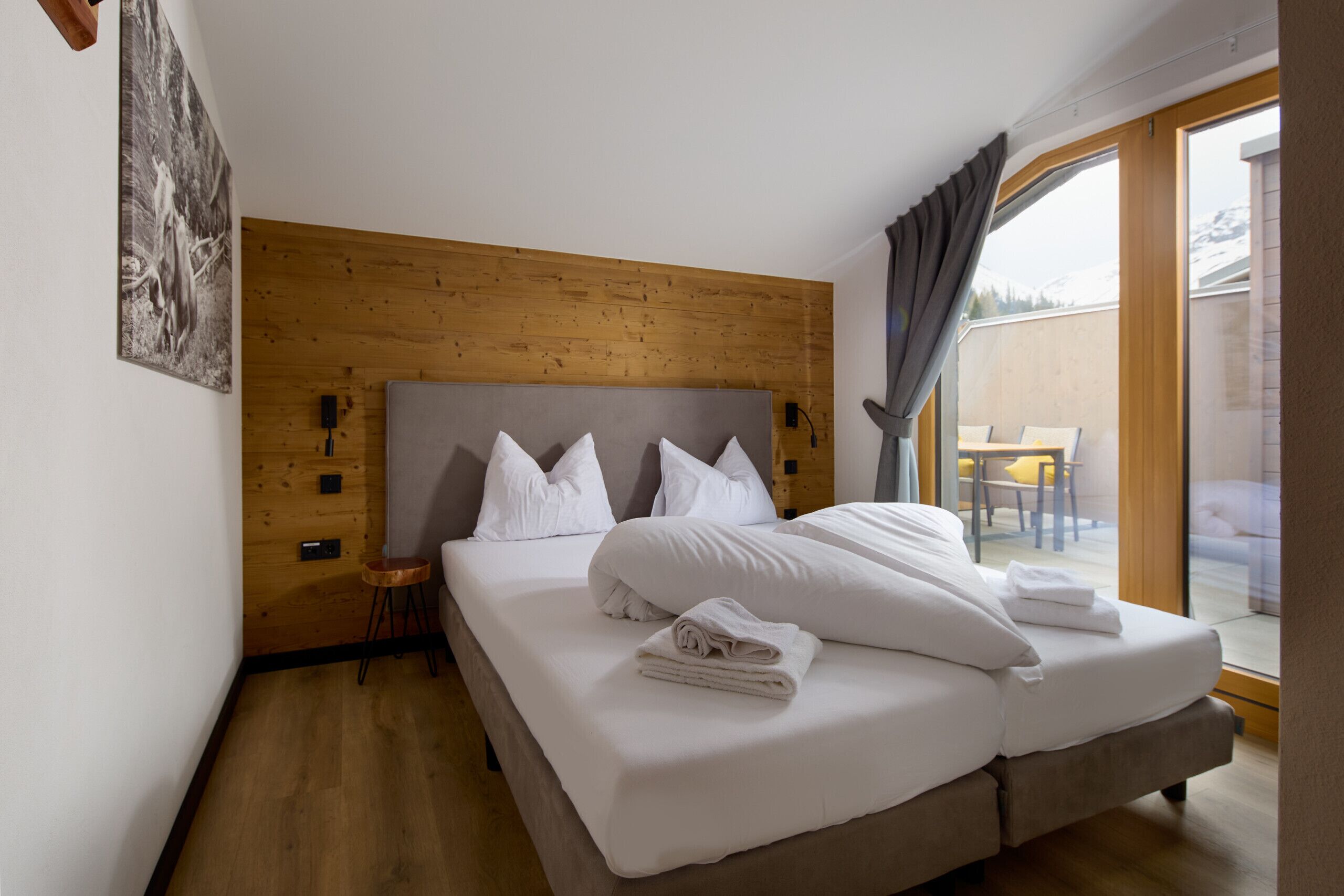 A bedroom with a wooden headboard, a large bed with white linens, a small stool, and a view of the outdoors through a glass door.