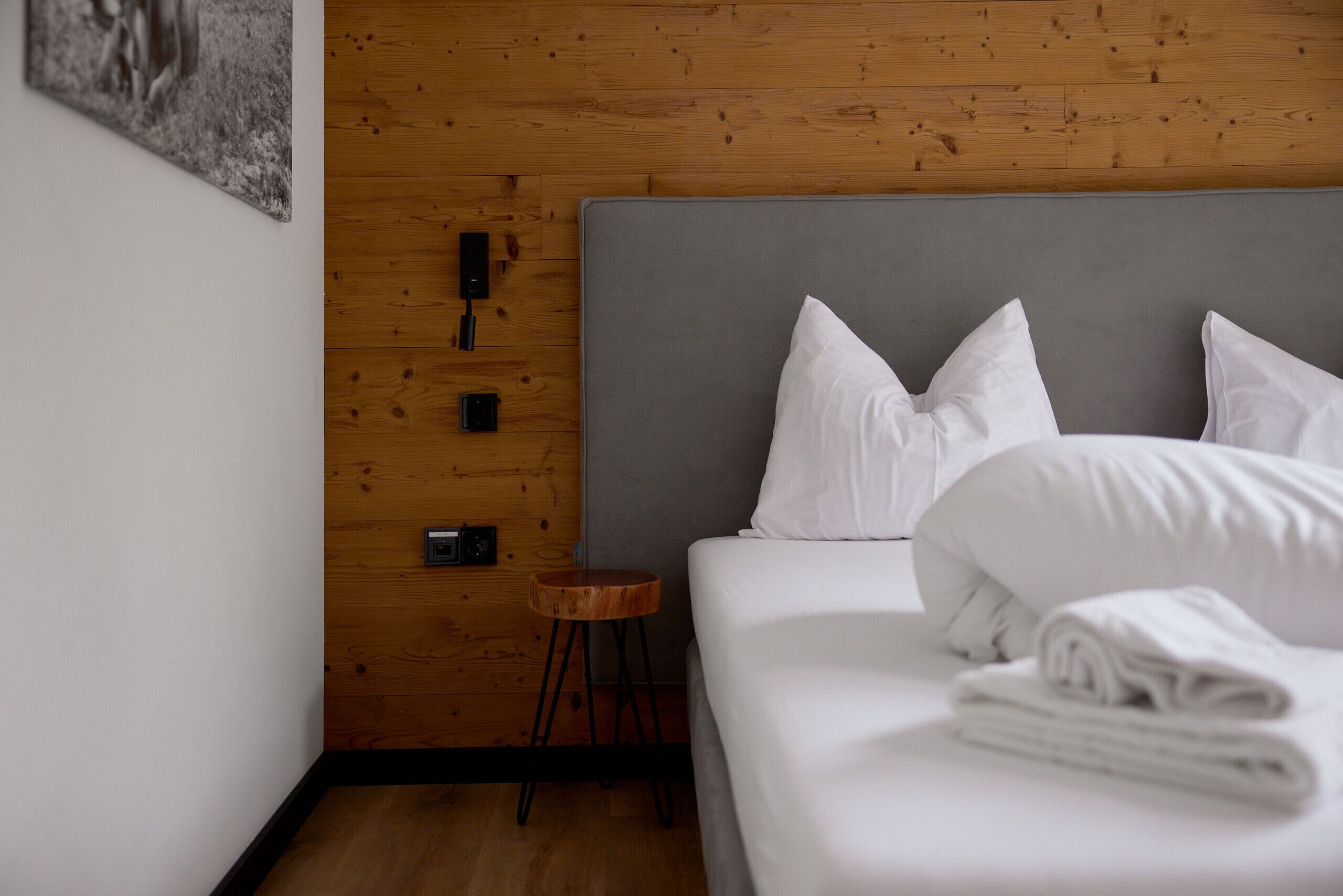 A neatly made bed with white linens and pillows, a small wooden stool, and a wall-mounted power outlet.