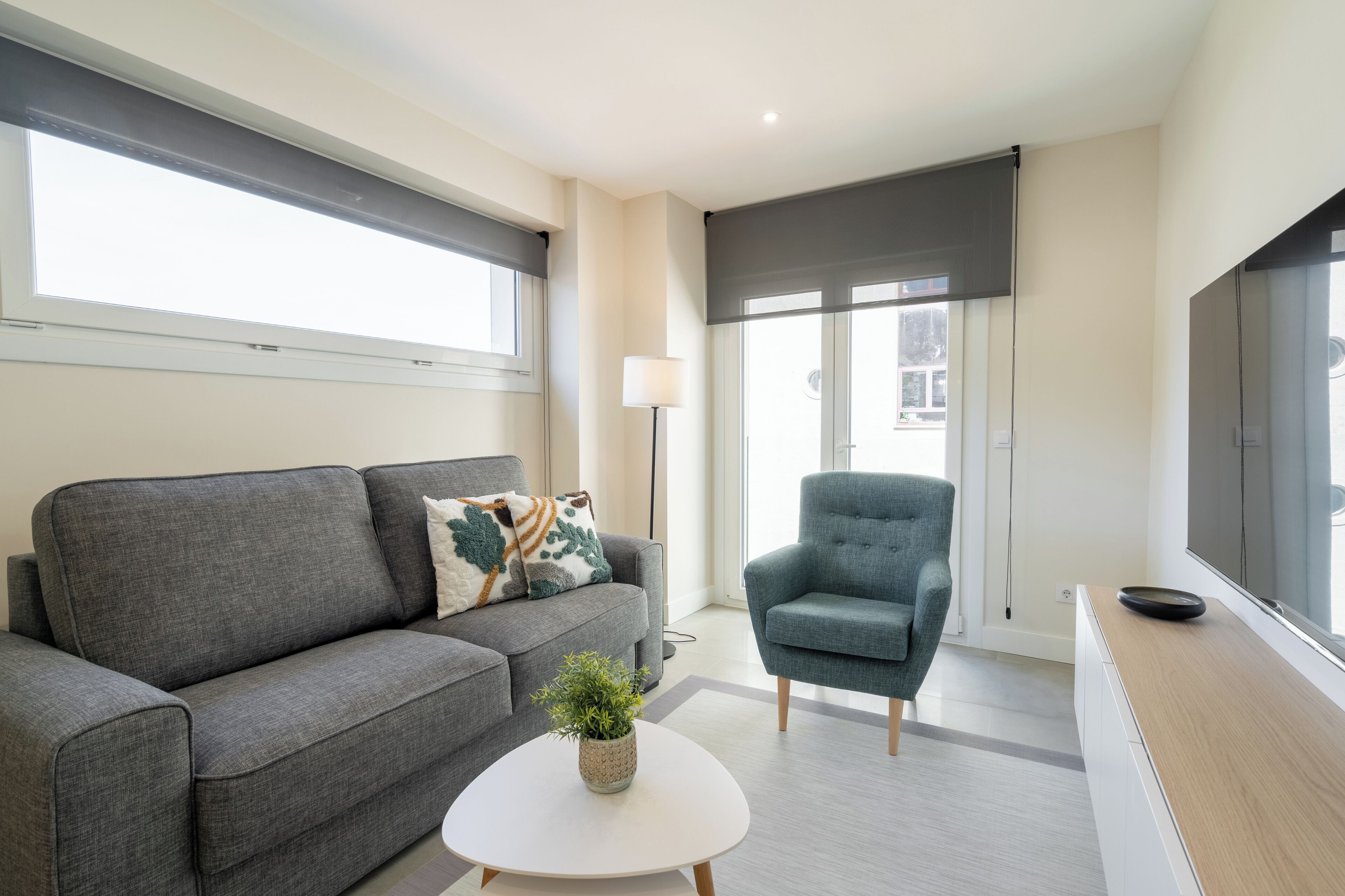 Ein modernes Wohnzimmer mit einem grauen Sofa, einem blauen Sessel, einem weißen Couchtisch mit Pflanze und einem Fernseher auf einem Holzboden.