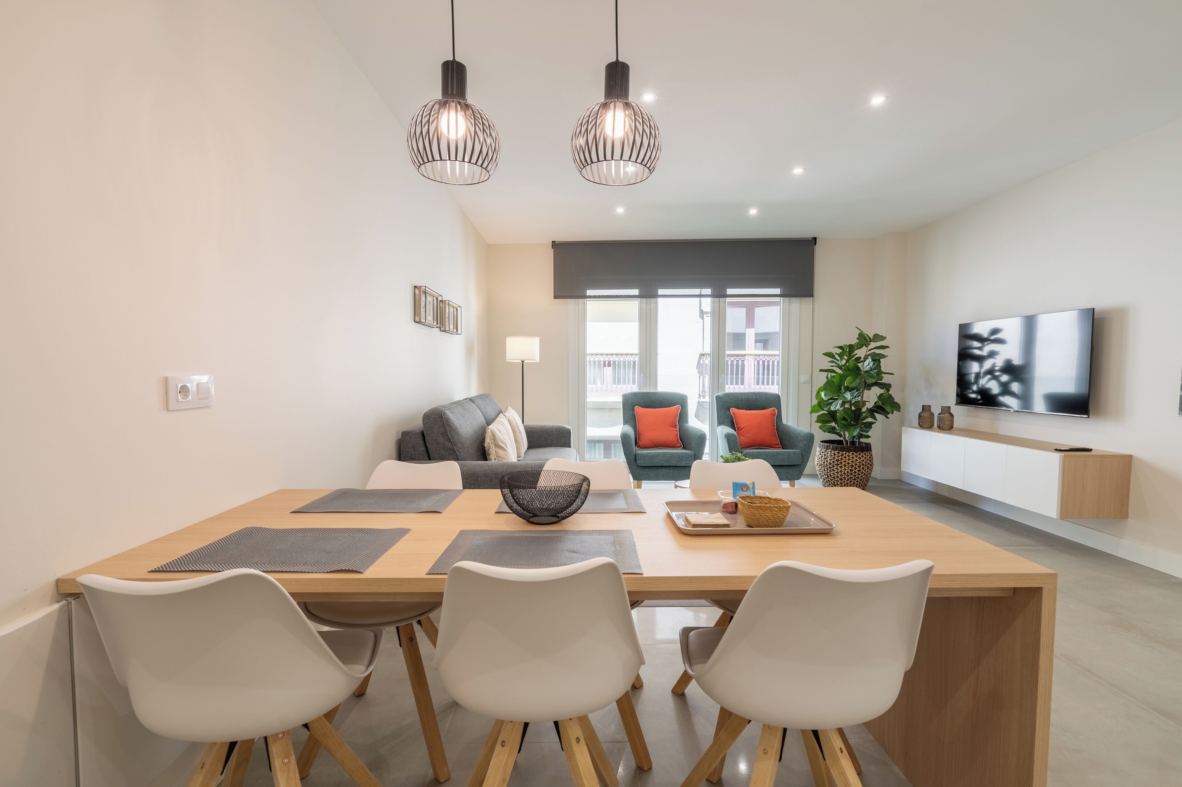 A modern living room with a wooden dining table, white chairs, a sofa, and a TV mounted on the wall.