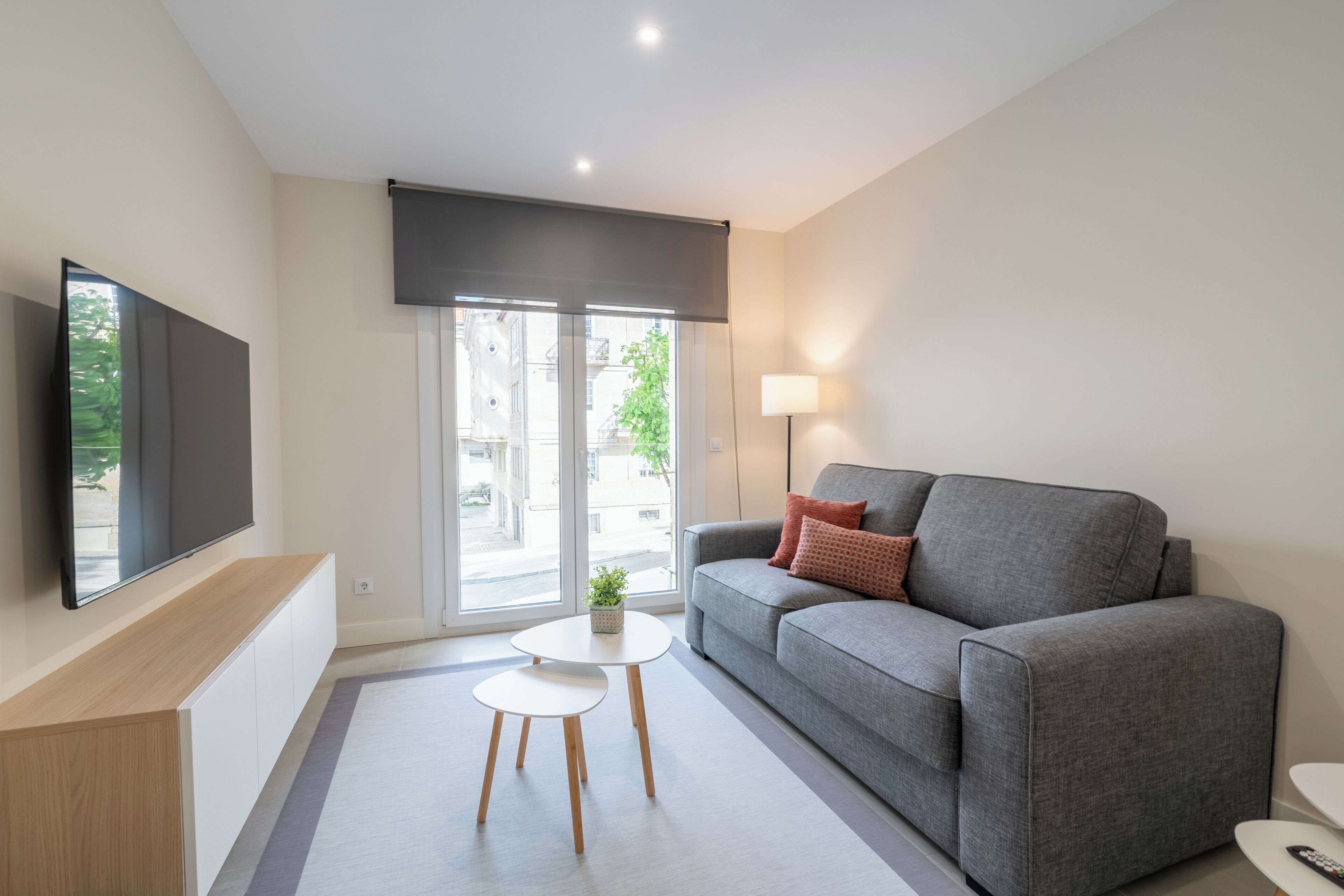 Ein modernes Wohnzimmer mit einem grauen Sofa, einem an der Wand montierten Flachbildfernseher, einem kleinen weißen Tisch und einer Stehlampe.