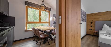 A modern kitchen with a dining area, wooden flooring, and a large window with a view of trees.