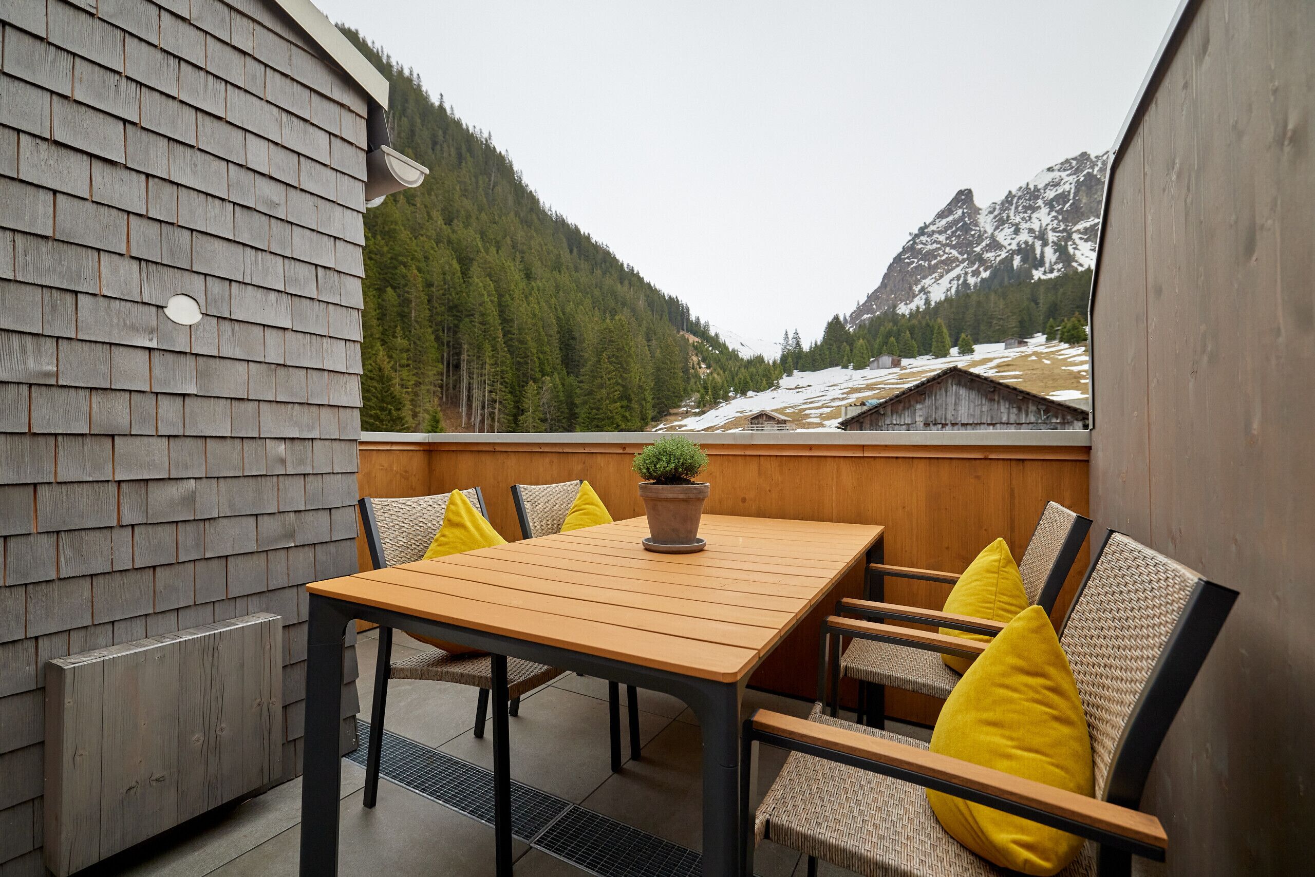 A wooden table with six chairs, a potted plant, and a view of snow-capped mountains and forested hills.