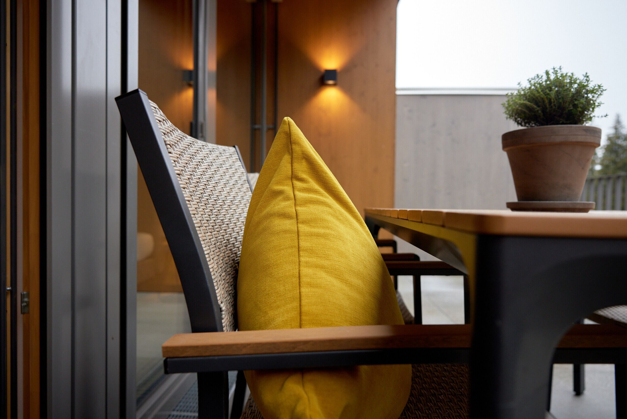 Un espace extérieur moderne, aménagé avec une table et des chaises en bois, un coussin jaune et une plante en pot.