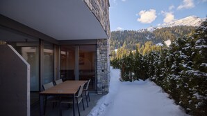 Un edificio moderno con fachada de piedra, una mesa y sillas de madera afuera y un paisaje montañoso nevado al fondo.