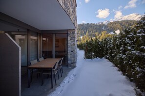 A modern building with a stone facade, a wooden table and chairs outside, and a snowy mountain landscape in the background.