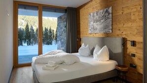 Un dormitorio moderno con una cama grande, vistas a un paisaje nevado a través de una puerta corredera, paneles de madera en las paredes y una obra de arte colgada.