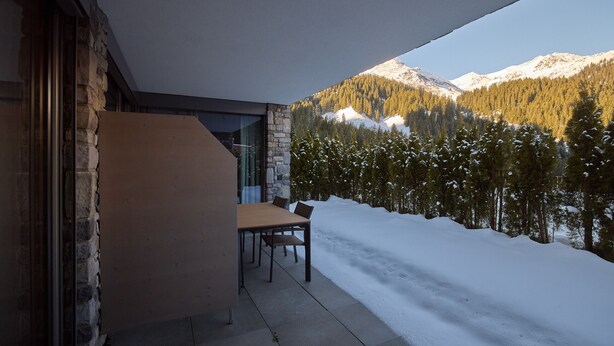 Un edificio moderno con fachada de piedra, una mesa y sillas de madera, y vistas a montañas y árboles cubiertos de nieve.