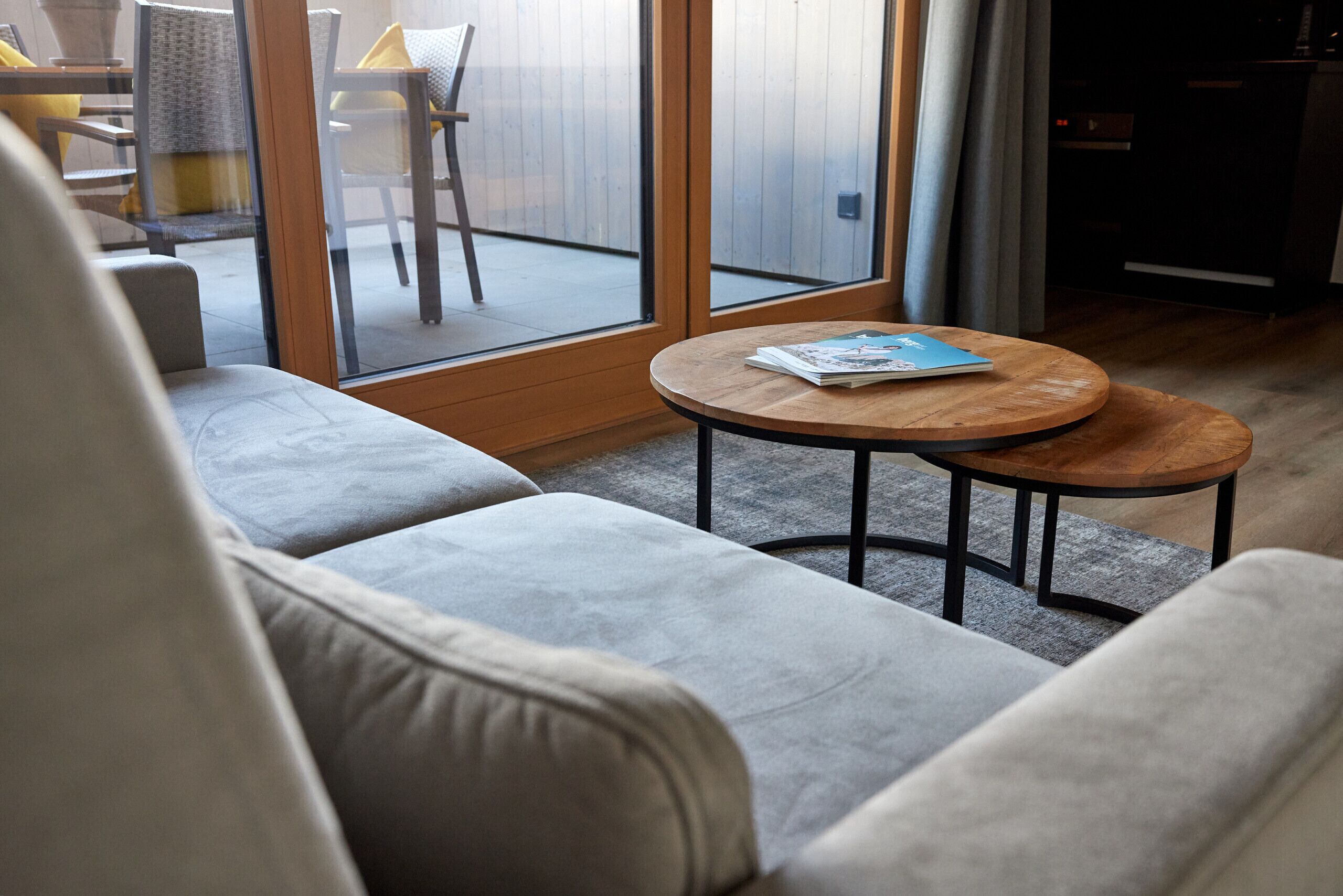 Ein modernes Wohnzimmer mit einer Couch, einem runden Holztisch und Blick durch Glastüren auf eine Außenpatio-Terrasse.