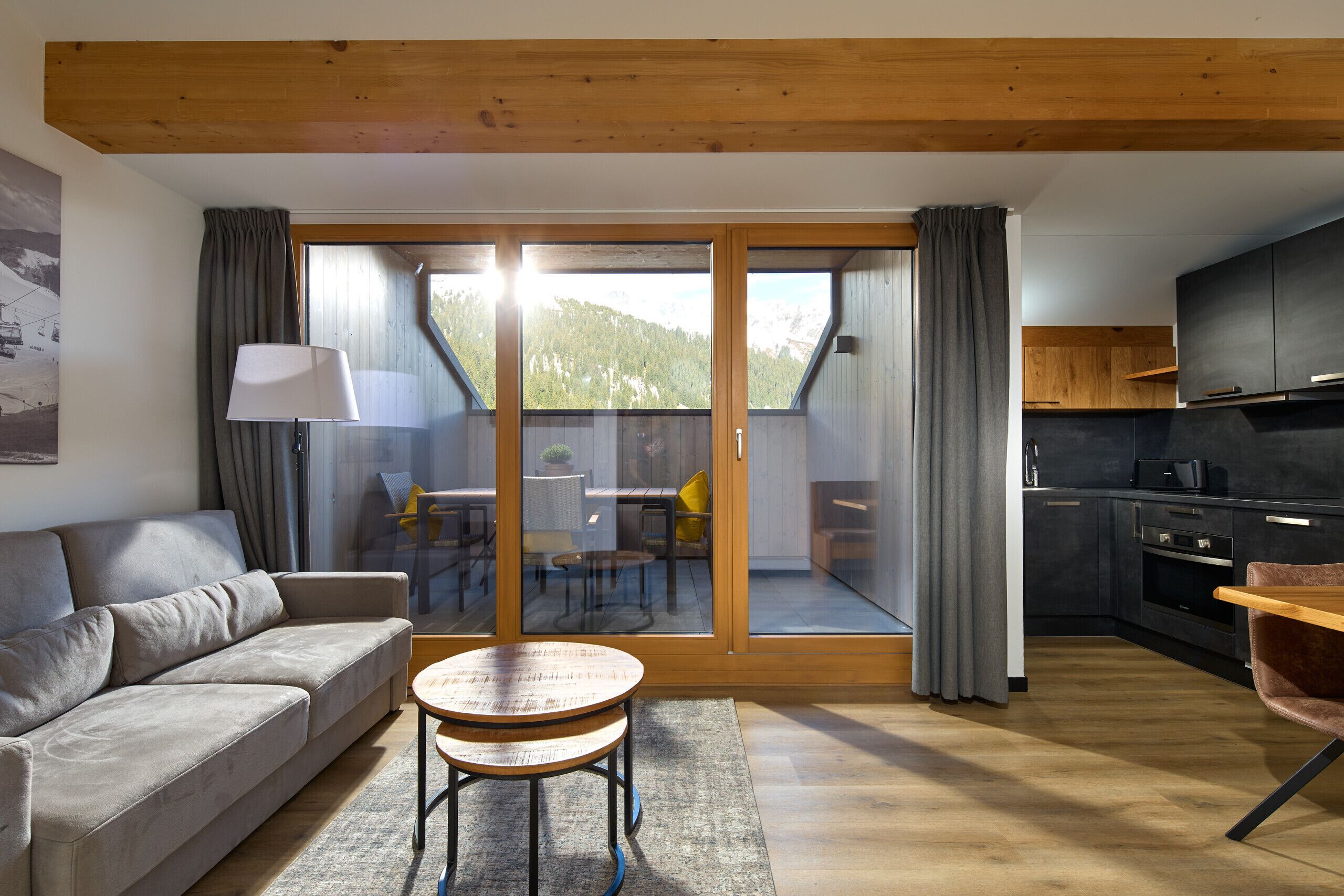 A modern living room with a sofa, a round wooden table, and a sliding glass door leading to a balcony with a mountain view.