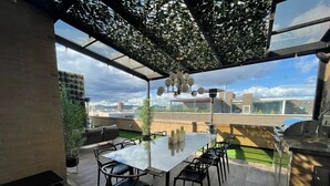 A rooftop terrace with a glass table, chairs, and a grill.