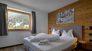A modern bedroom with a large bed, a wooden headboard, a window with curtains, and a black and white photograph on the wall.