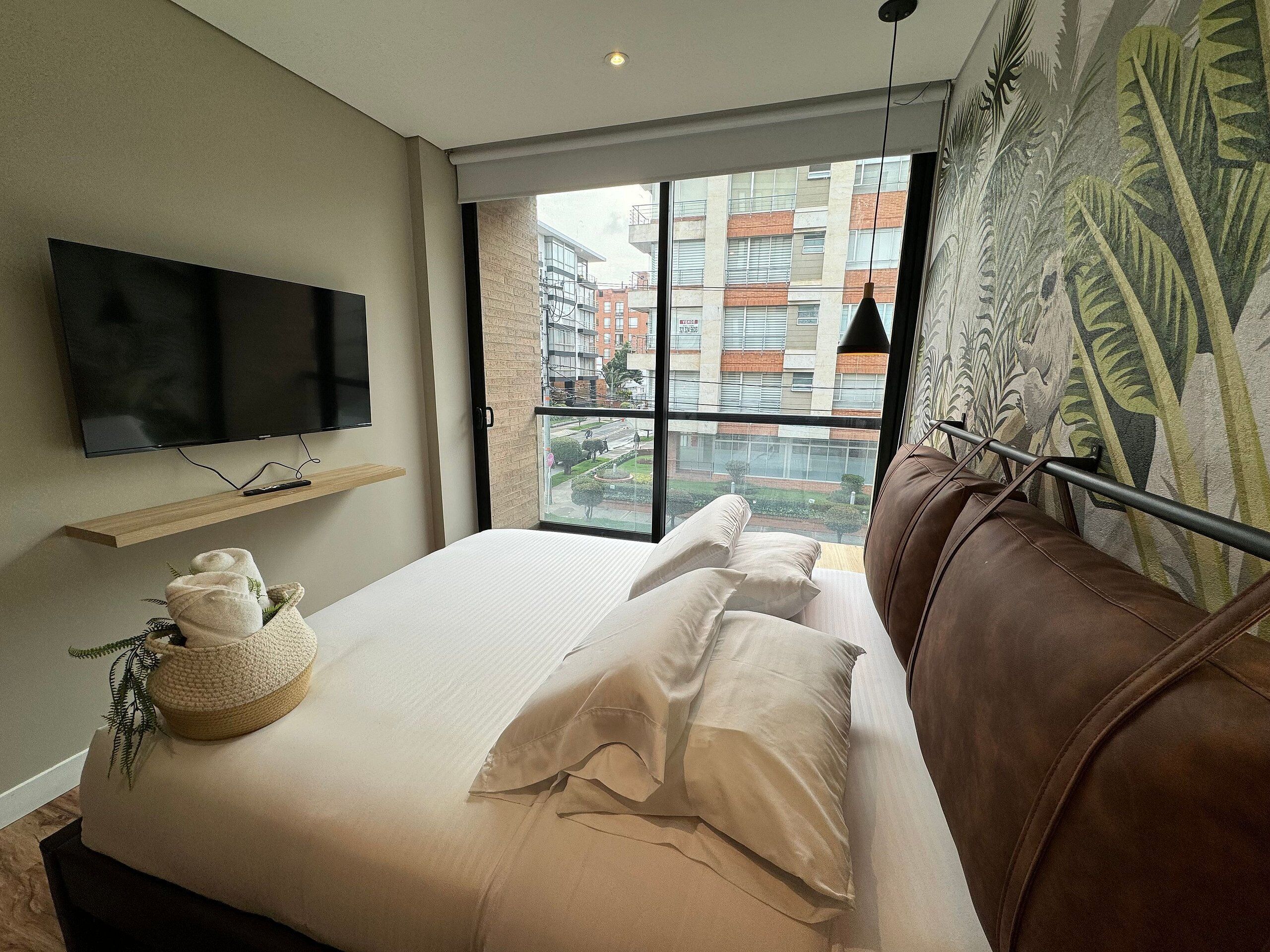A modern hotel room with a large bed, a mounted TV, and a view of the city through a large window.