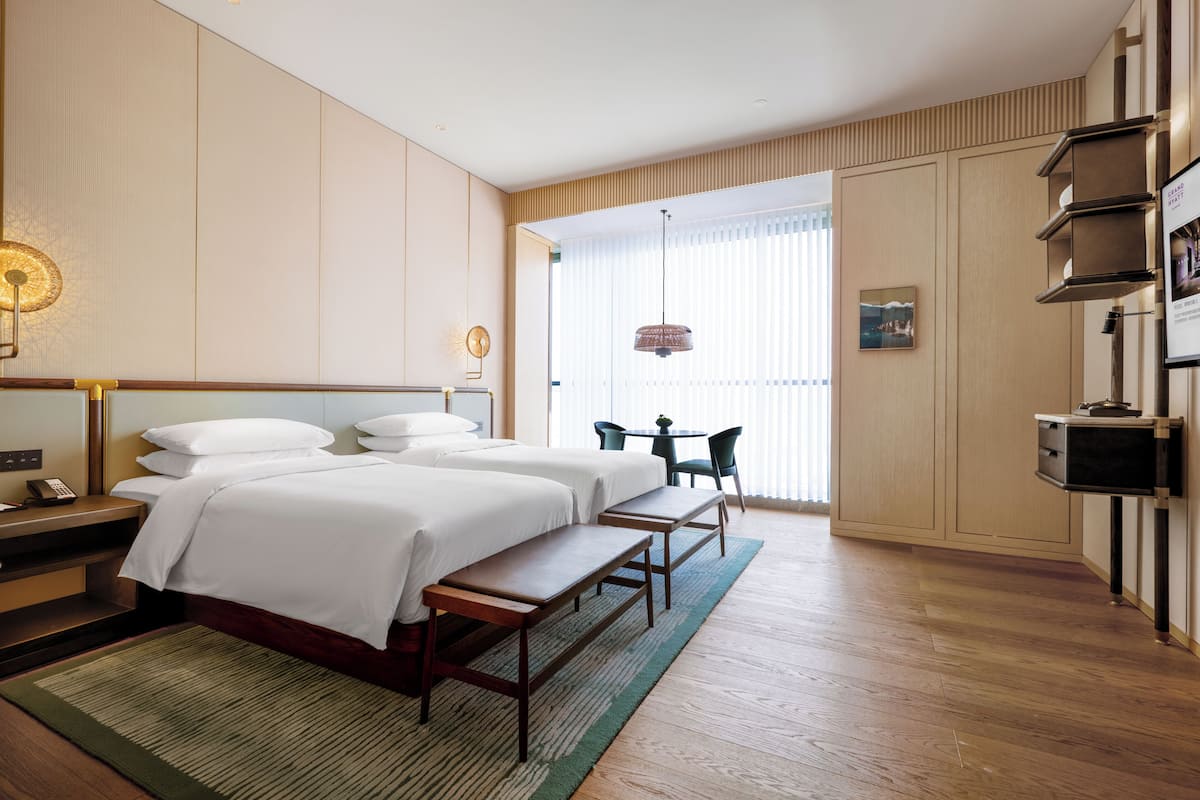 Quarto de hotel com duas camas, uma mesa, uma cadeira, uma mesinha de centro e uma janela grande com persianas.