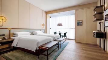 Une chambre d’hôtel avec deux lits, un bureau, une chaise, une petite table et une grande fenêtre avec des stores.