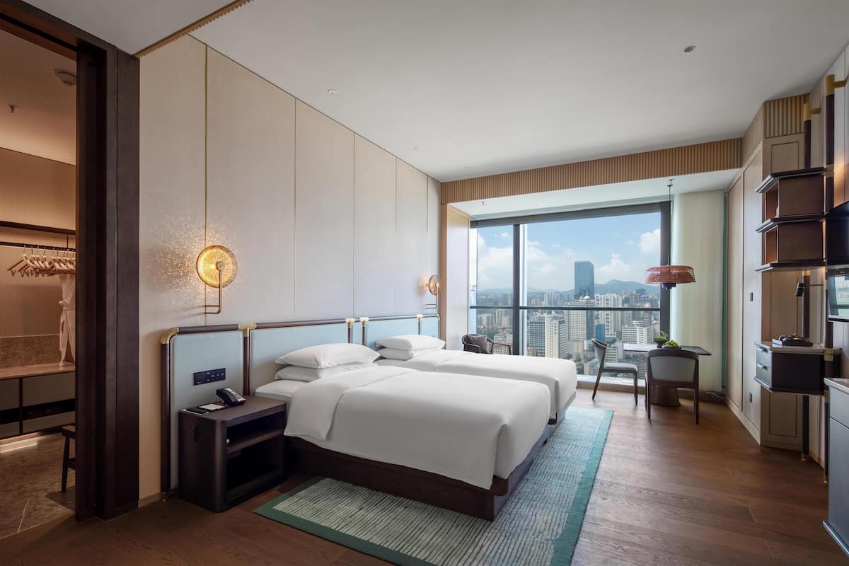 Quarto de hotel moderno com uma janela grande, duas camas, uma escrivaninha e vista para a cidade.