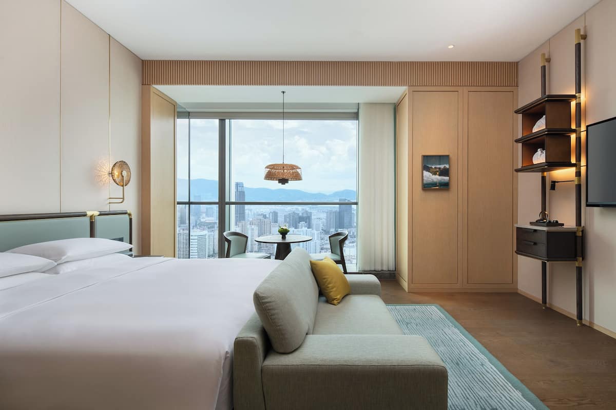 Quarto de hotel com uma cama grande, um sofá, uma mesa de jantar com cadeiras, uma TV e vista para a cidade.