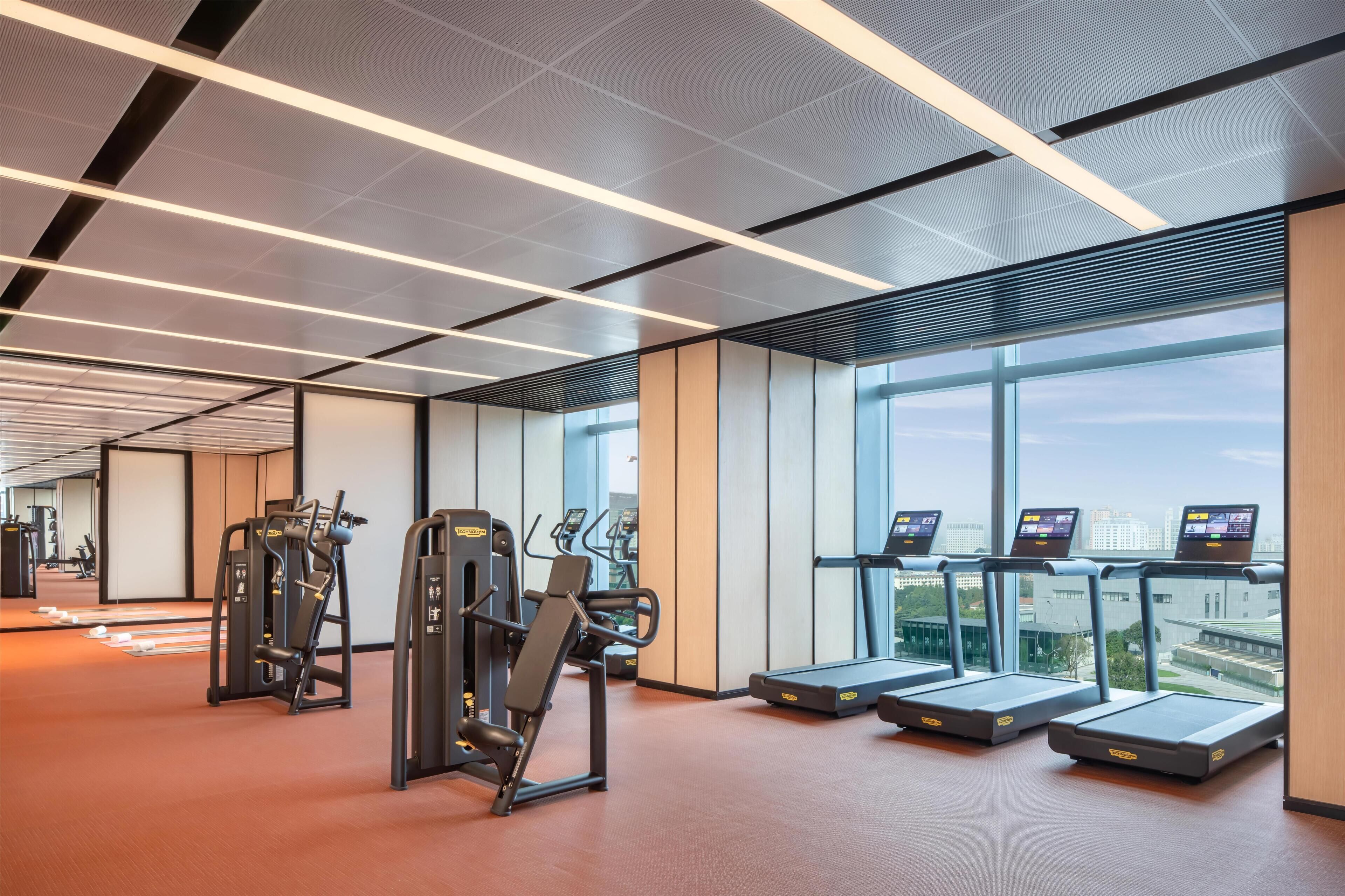 Un gimnasio con equipo de ejercicio, cintas de correr y grandes ventanales con vistas a la ciudad.