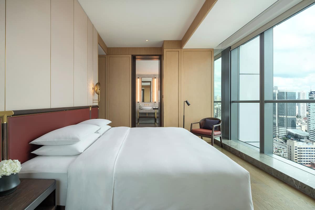 Quarto de hotel moderno com uma cama grande, vista para a cidade e uma mesa com cadeira.