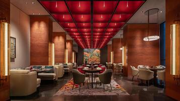 A modern hotel lobby with a red ceiling, multiple dining tables, and comfortable seating.