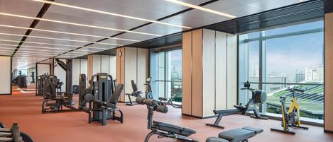 Un gimnasio con diverso equipo de ejercicio, amplias ventanas y vistas al paisaje urbano.