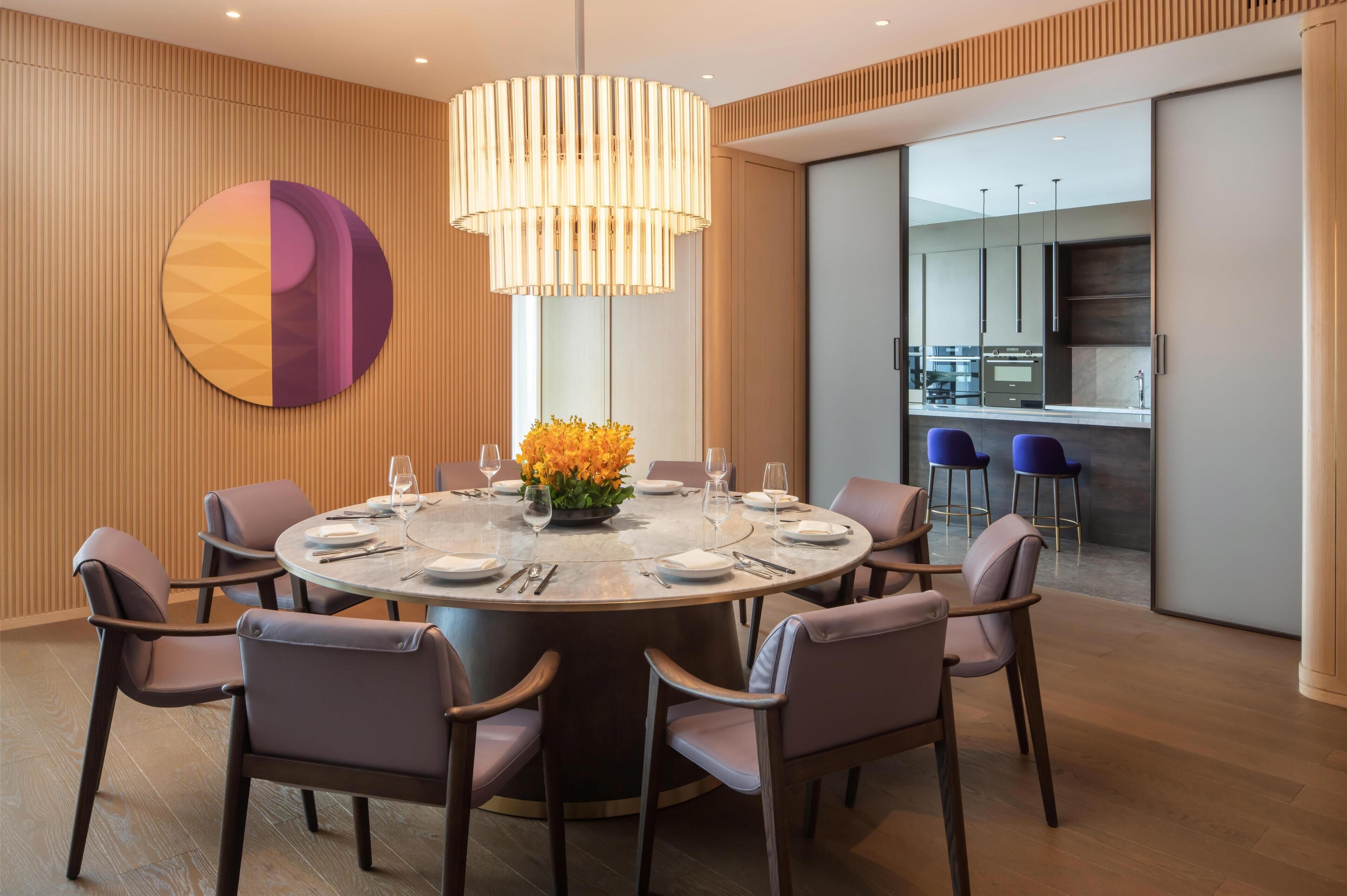 A modern dining area with a round table, chairs, a large pendant light, and a decorative wall piece.