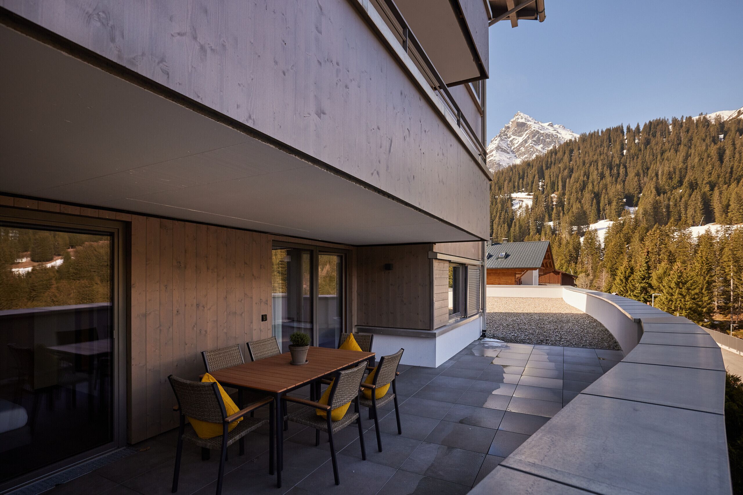 An outdoor dining area with a wooden table and chairs, a potted plant, and a view of a mountain range.