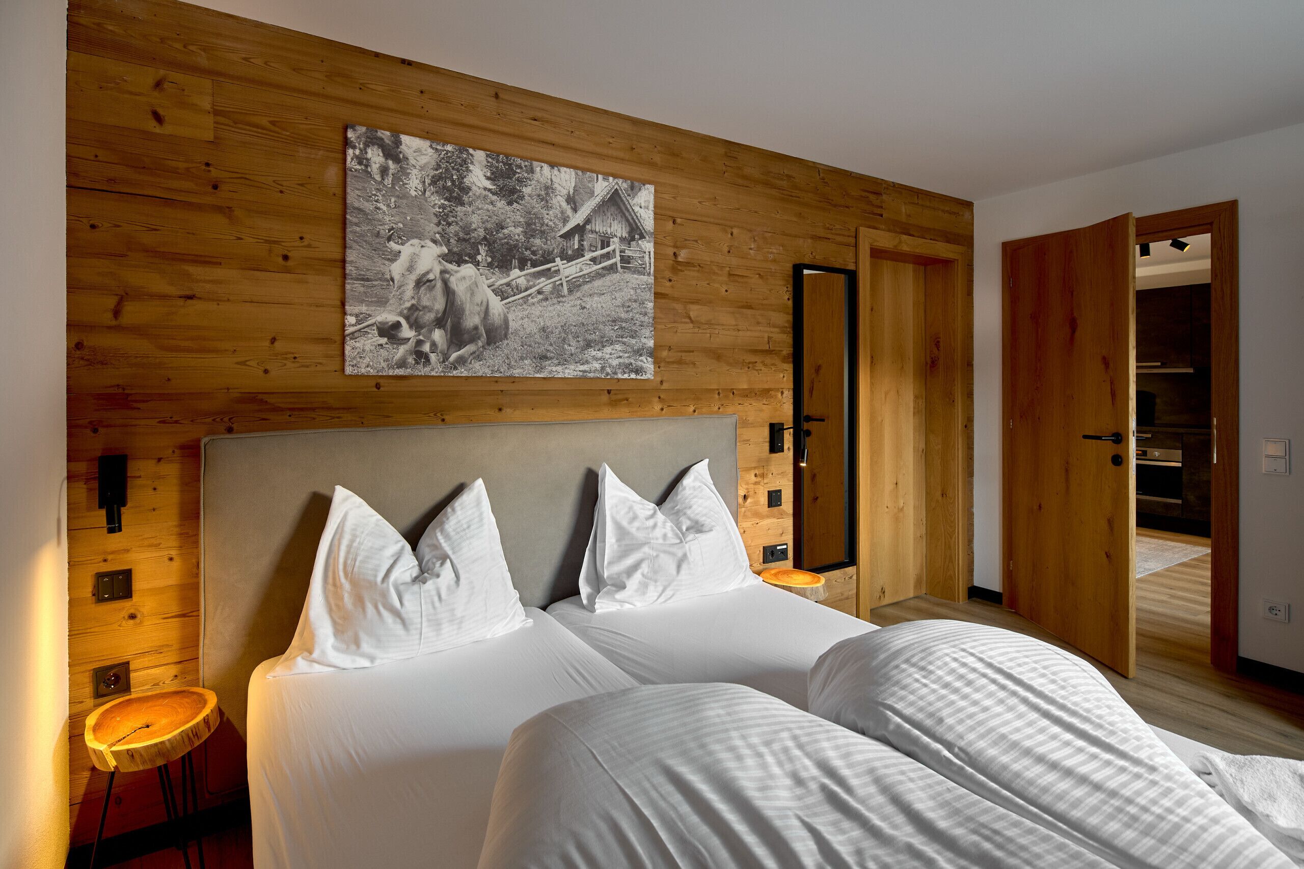 Un dormitorio con cabecera de madera, una cama grande con ropa de cama blanca, una mesita redonda pequeña y una puerta de madera que da a otra habitación.