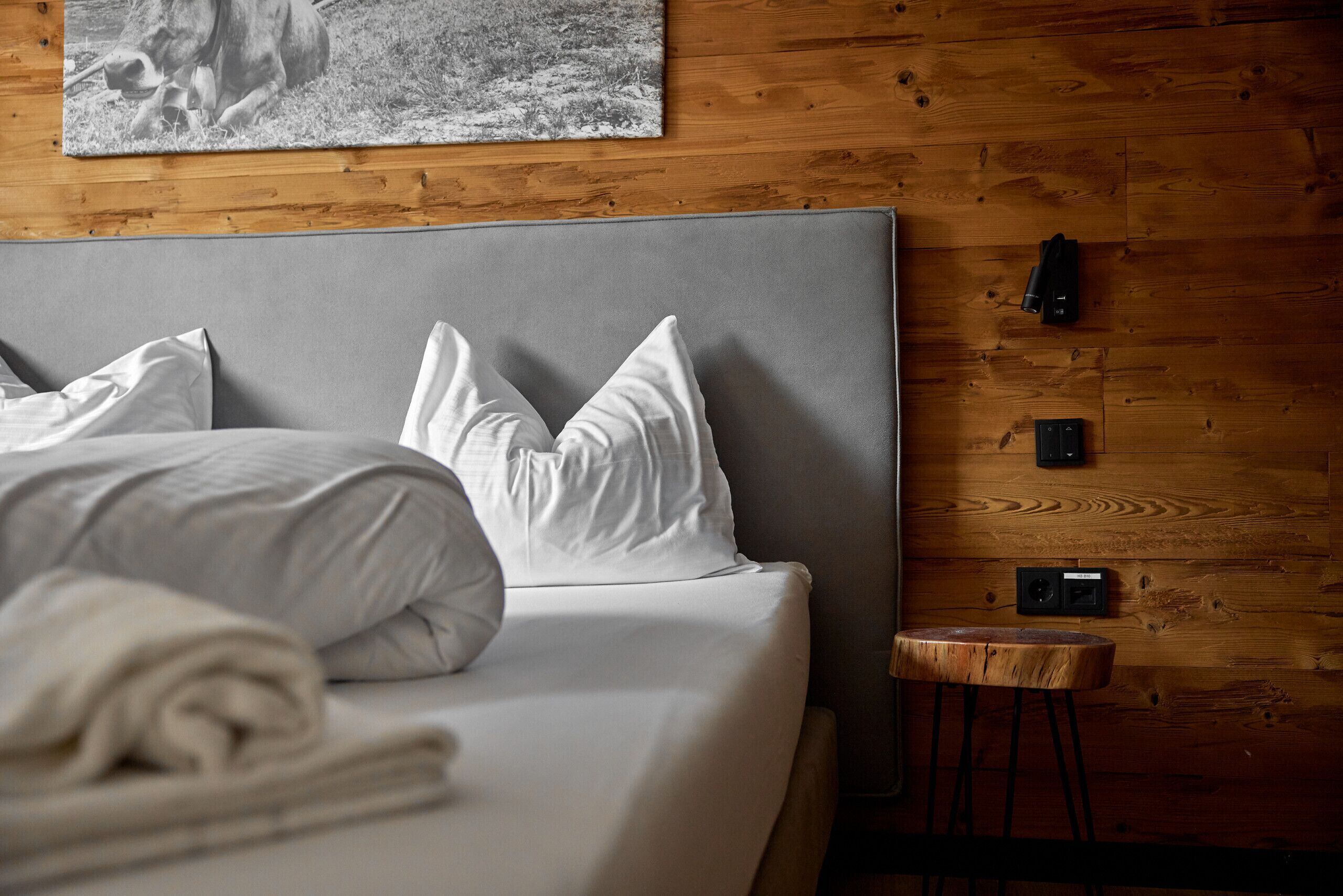 Un lit bien fait, avec du linge de lit blanc et des oreillers, un mur en bois orné d’un tableau, et un petit tabouret en bois.