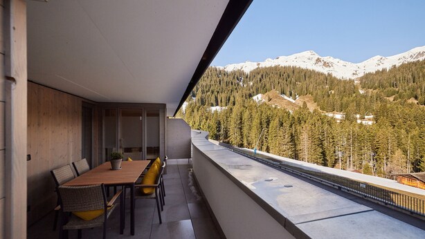 Un balcón con mesa y sillas de madera, con vista a un paisaje montañoso de picos nevados y densos bosques.