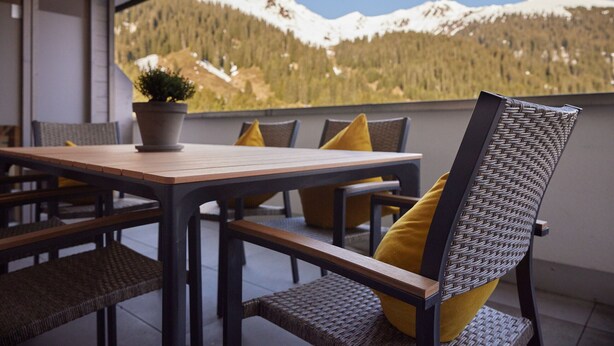 A balcony with a wooden table and wicker chairs, a potted plant, and a mountain view.