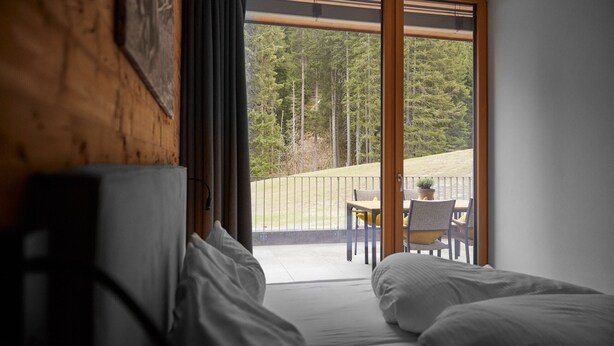Une chambre dotée d’une grande porte-fenêtre coulissante donnant sur un balcon aménagé avec une table et des chaises, et offrant une vue sur une zone boisée.