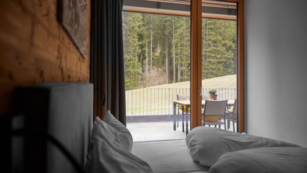 Une chambre dotée d’une grande porte-fenêtre coulissante donnant sur un balcon aménagé avec une table et des chaises, et offrant une vue sur une zone boisée.