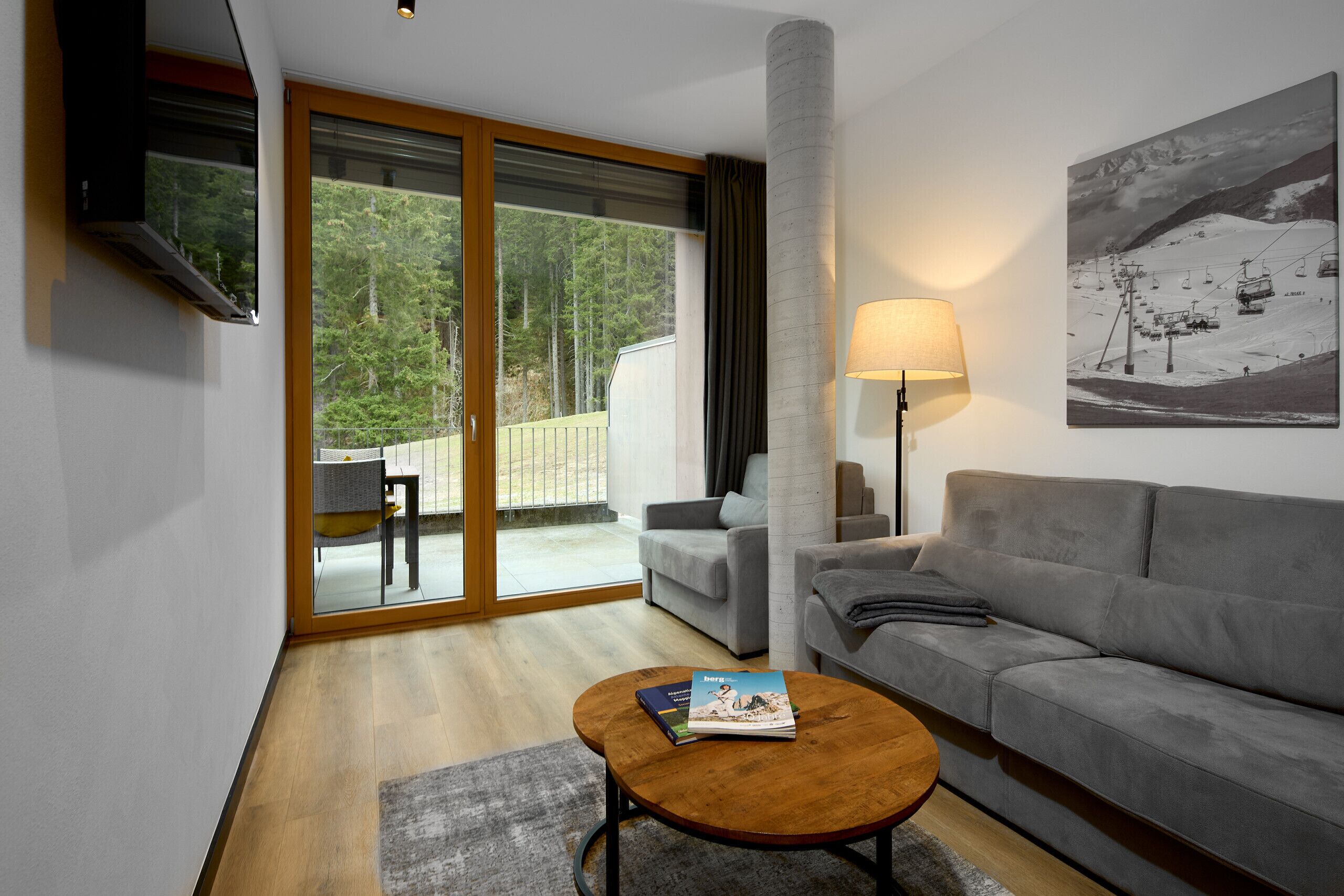 A modern living room with a grey sofa, a wooden coffee table with magazines, a floor lamp, and a large window with a view of a snowy mountain and forest.