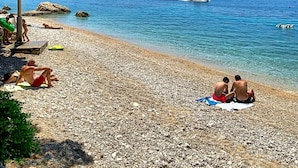 Une plage de galets avec une eau d’un bleu limpide, un bateau et des gens qui prennent un bain de soleil.