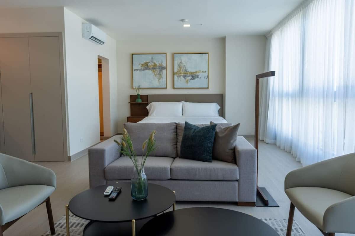 Een moderne hotelkamer met een grijze bank, een ronde zwarte salontafel en een bed met witte lakens. Boven het bed hangen twee kunstwerken in lijsten en er is een raam met transparante gordijnen.