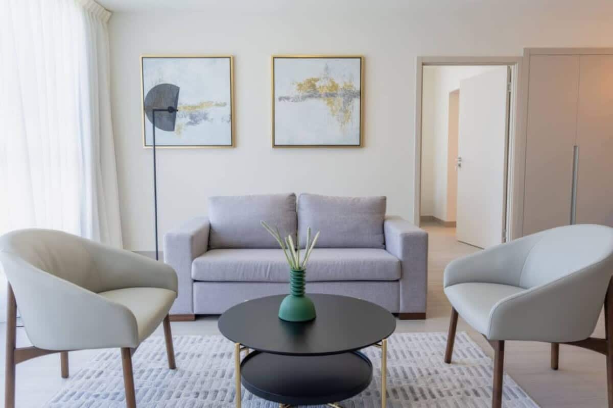 Een moderne woonkamer met een grijze bank, twee fauteuils, een ronde zwarte salontafel en twee abstracte schilderijen aan de muur.