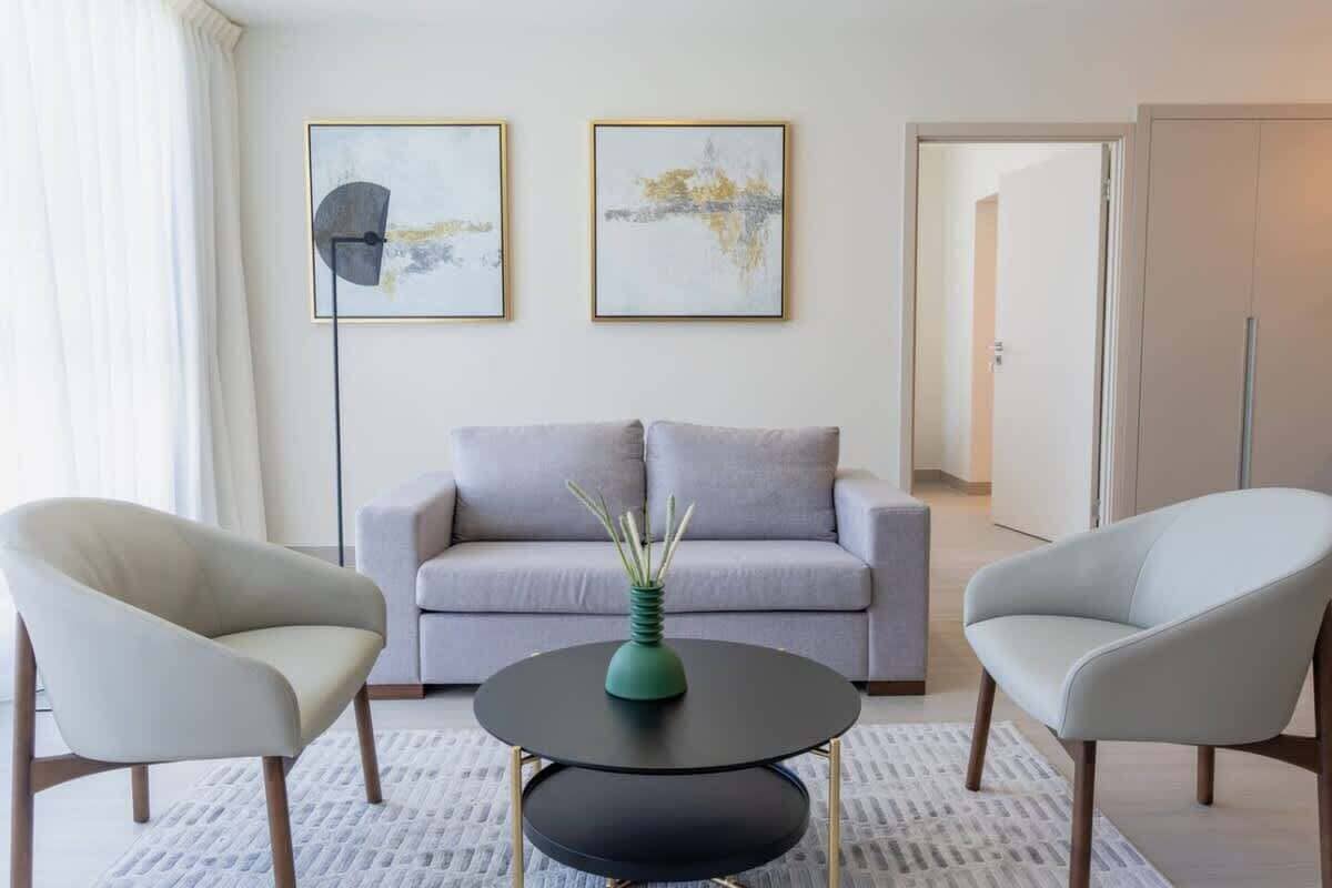 Una sala de estar moderna con un sofá gris, dos sillones, una mesita redonda y dos cuadros abstractos en la pared.