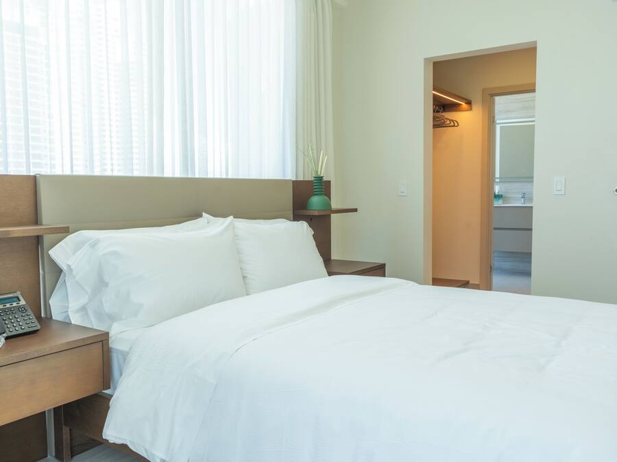 Habitación de hotel con cama, teléfono junto a la cama, mesita de noche, ventana con cortinas y un espejo en la habitación contigua.