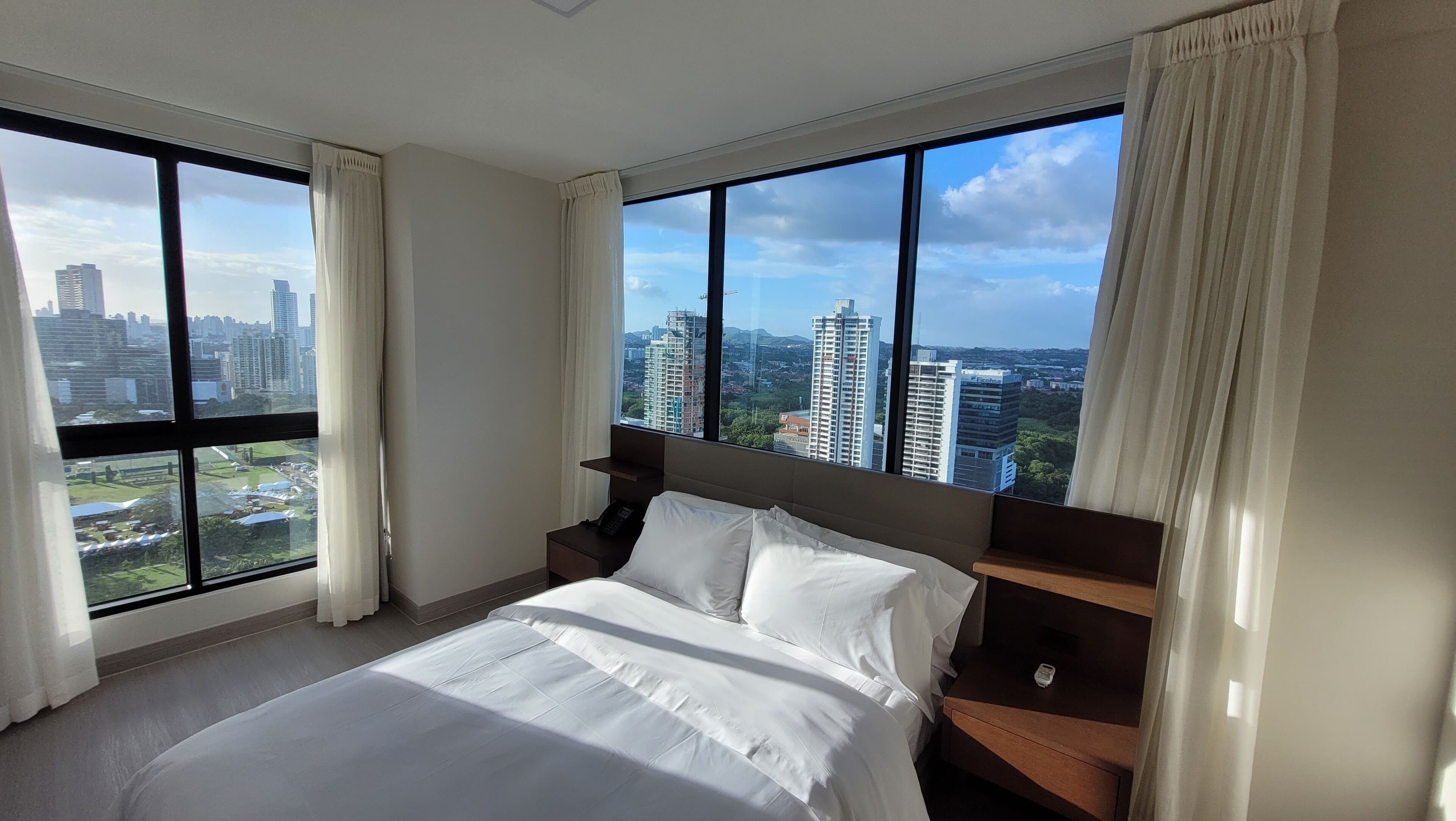 Habitación de hotel con una cama grande, una mesita de noche y vistas a un paisaje urbano a través de amplias ventanas.