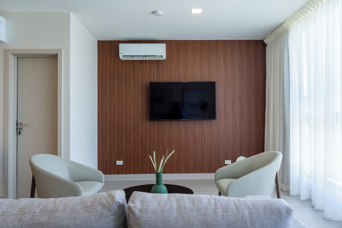 Een moderne woonkamer met een flatscreen televisie aan een houten wand, twee fauteuils, een salontafel en een deur aan de linkerkant.