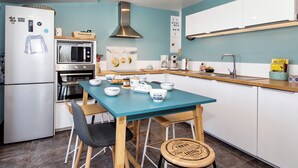 A modern kitchen with a teal countertop, stainless steel appliances, and a wooden dining table with chairs. - Maison 3 Bord de Mer, Chez Ty Suzanne à 150m des Plages de Sable fin (Cléder)