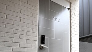 Une porte moderne dotée d’une serrure numérique et portant le numéro 202, située dans une pièce aux murs en briques blanches et au sol gris.